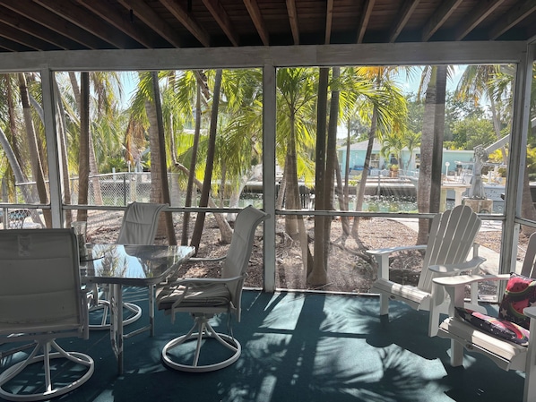 A covered outdoor seating area with a table and chairs, surrounded by palm trees and a view of a marina.