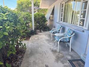 A covered patio with two white chairs and a small table, surrounded by greenery and a building with windows.