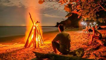 A beach scene with a bonfire, a person sitting by the fire, and a group of people relaxing on the sand.