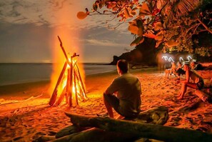 A beach scene with a bonfire, a person sitting by the fire, and a group of people relaxing on the sand.