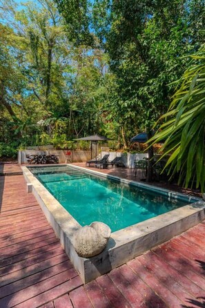 A rectangular swimming pool with a stone edge, surrounded by a wooden deck and lush greenery.