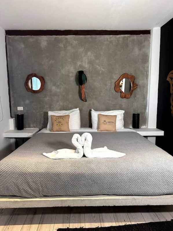 A bed with a grey textured bedspread, two pillows with a tropical design, and two white swan-shaped towels.