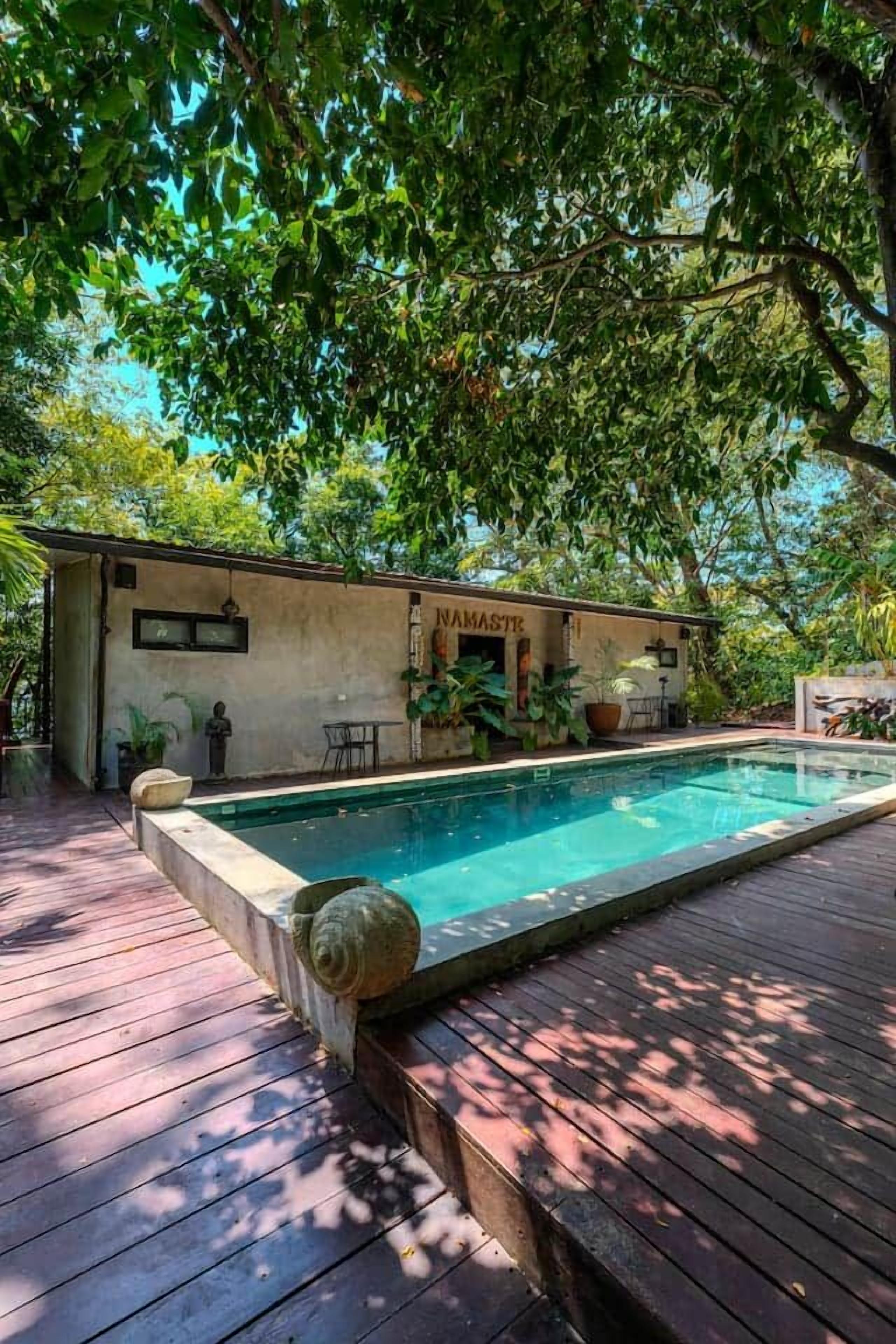 A swimming pool with a wooden deck, surrounded by lush greenery and a building with a sign that reads 'NAMASTR'.