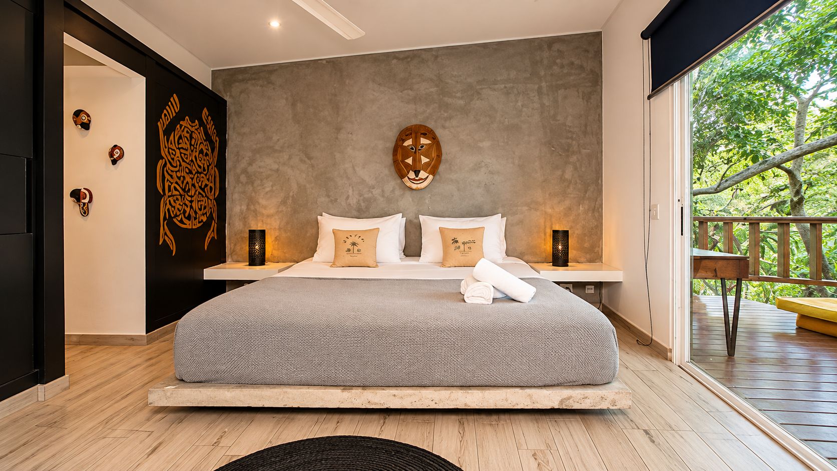 A modern bedroom with a large bed, a concrete wall with a wooden mask, and a balcony with a wooden railing.