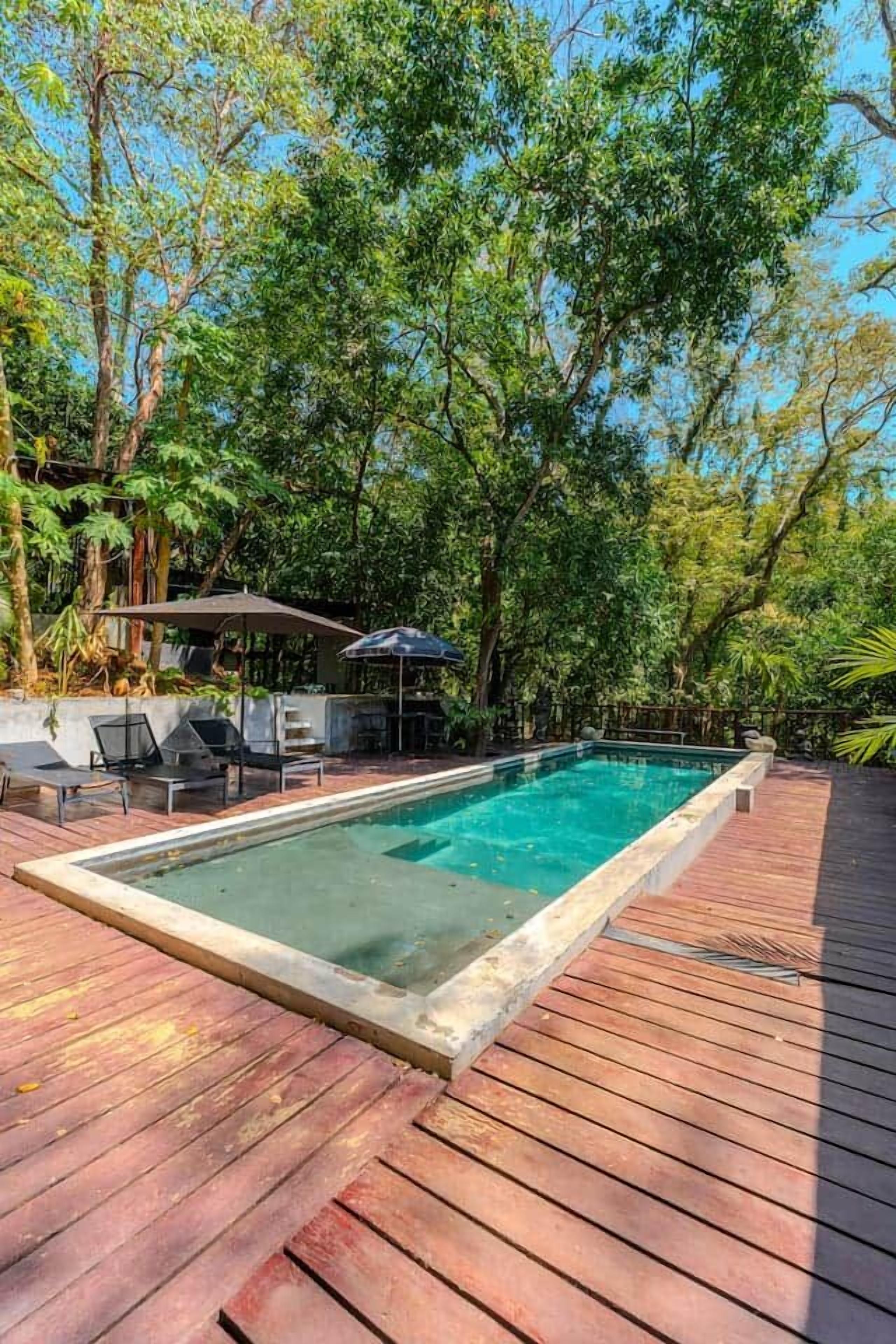 A rectangular swimming pool with a wooden deck surrounding it, set in a lush green environment.