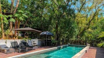 A rectangular swimming pool with a wooden deck surrounding it, set in a lush green environment.