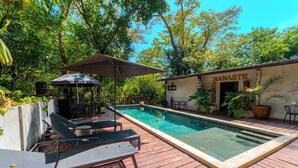 Una piscina con sillas de descanso y una sombrilla, rodeada de vegetación y un edificio con la leyenda 'MAMASTE'.