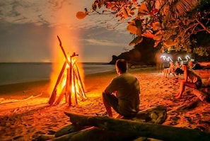 A beach scene with a bonfire, a person sitting by the fire, and a group of people relaxing on the sand.