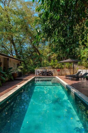 A rectangular swimming pool with a wooden deck surrounding it, a covered seating area, and lush greenery in the background.