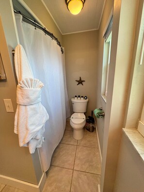 A narrow bathroom with a shower curtain, a toilet, a star decoration on the wall, and a trash can. - The Cape Escape | Cape Coral (Cape Coral)