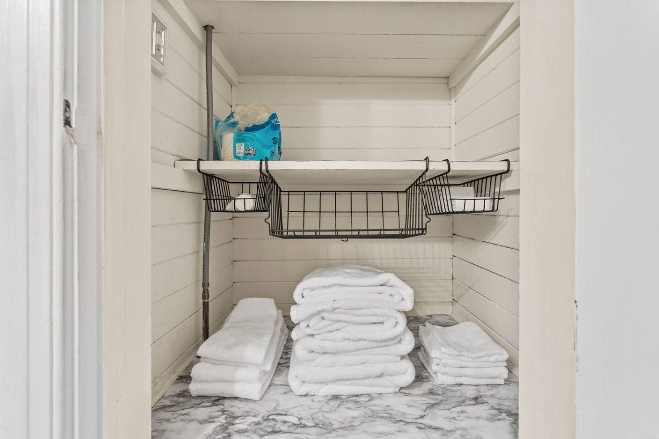 Une petite salle de bain aux murs blancs, dotée d’un sol en marbre, d’un porte-serviettes et d’une étagère sur laquelle se trouve un sac contenant des articles de toilette.