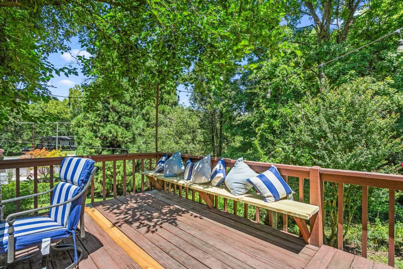 A wooden deck with a bench and a chair, surrounded by lush greenery.