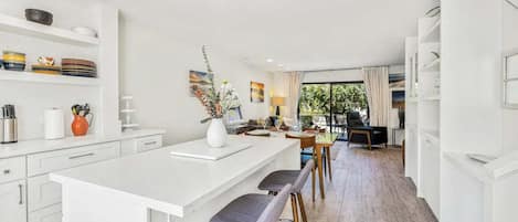 A modern kitchen with white cabinetry, a central island with bar stools, and a dining area with a view through large windows.