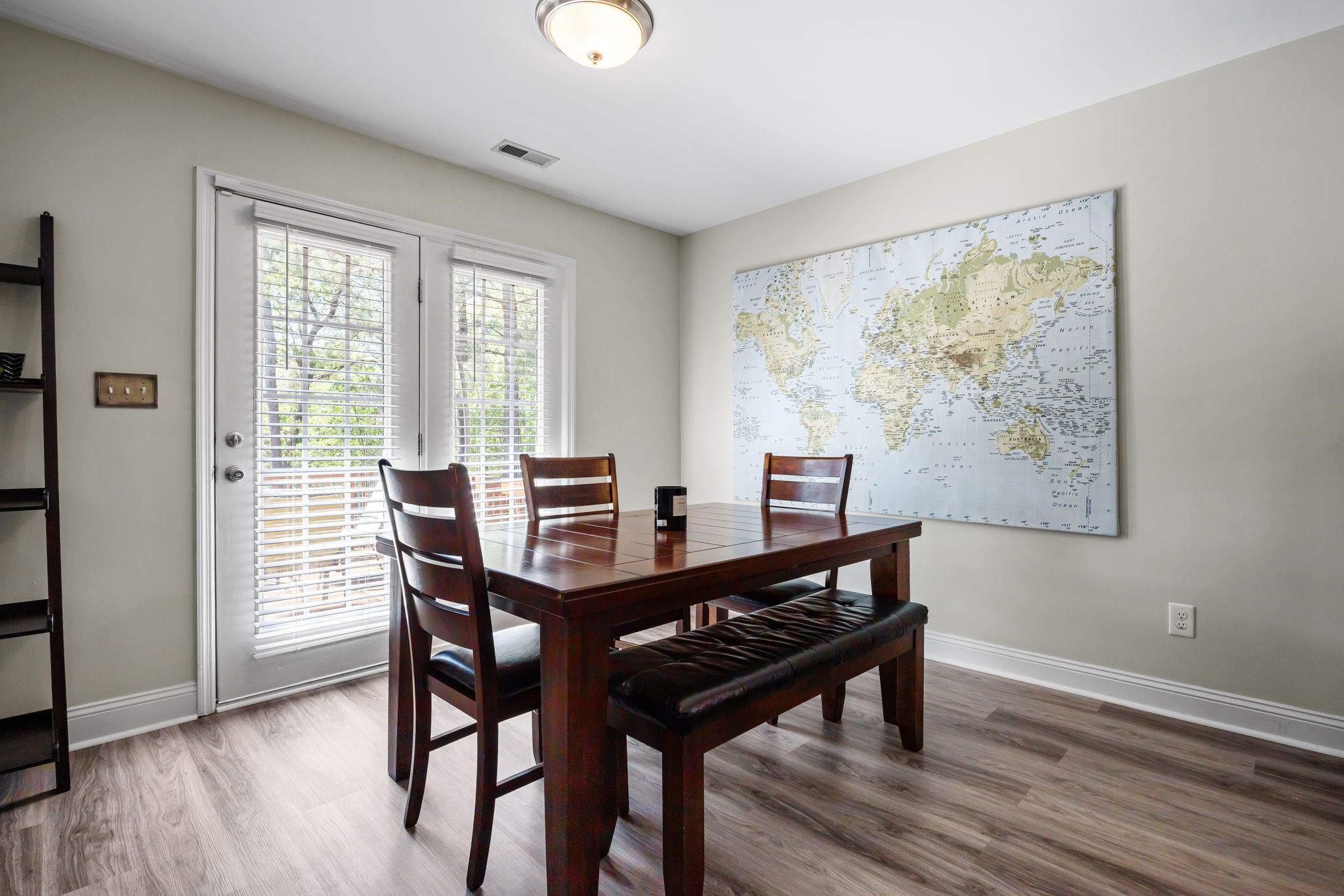A dining room with a wooden table, four chairs, a bench, a large world map on the wall, and a sliding glass door.