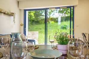 A dining table set with various glassware and a bowl, a potted plant with flowers, and a view of a green garden through a glass door. - La Vague Douce - Mer & Pinède, plage accès à pied (Douarnenez)