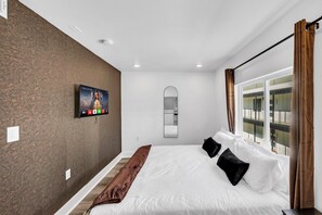 Una habitación de hotel con cama, televisor montado en la pared, un ventanal con cortinas y un espejo.