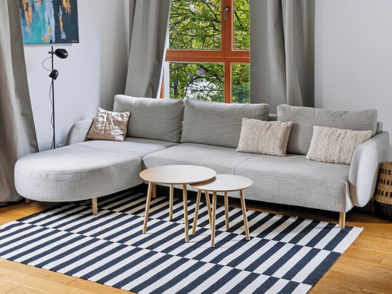 Een moderne woonkamer met een grijze modulaire bank, twee ronde houten tafels, een zwart-wit geruit tapijt en een raam met uitzicht op bomen.