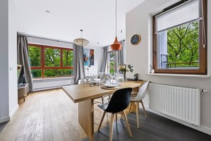 A dining area with a wooden table, chairs, and a vase of flowers. There are windows with curtains, a clock on the wall, and a radiator. - Warsaw Green Żoliborz Apt — Parking & Balcony (Warszawa)