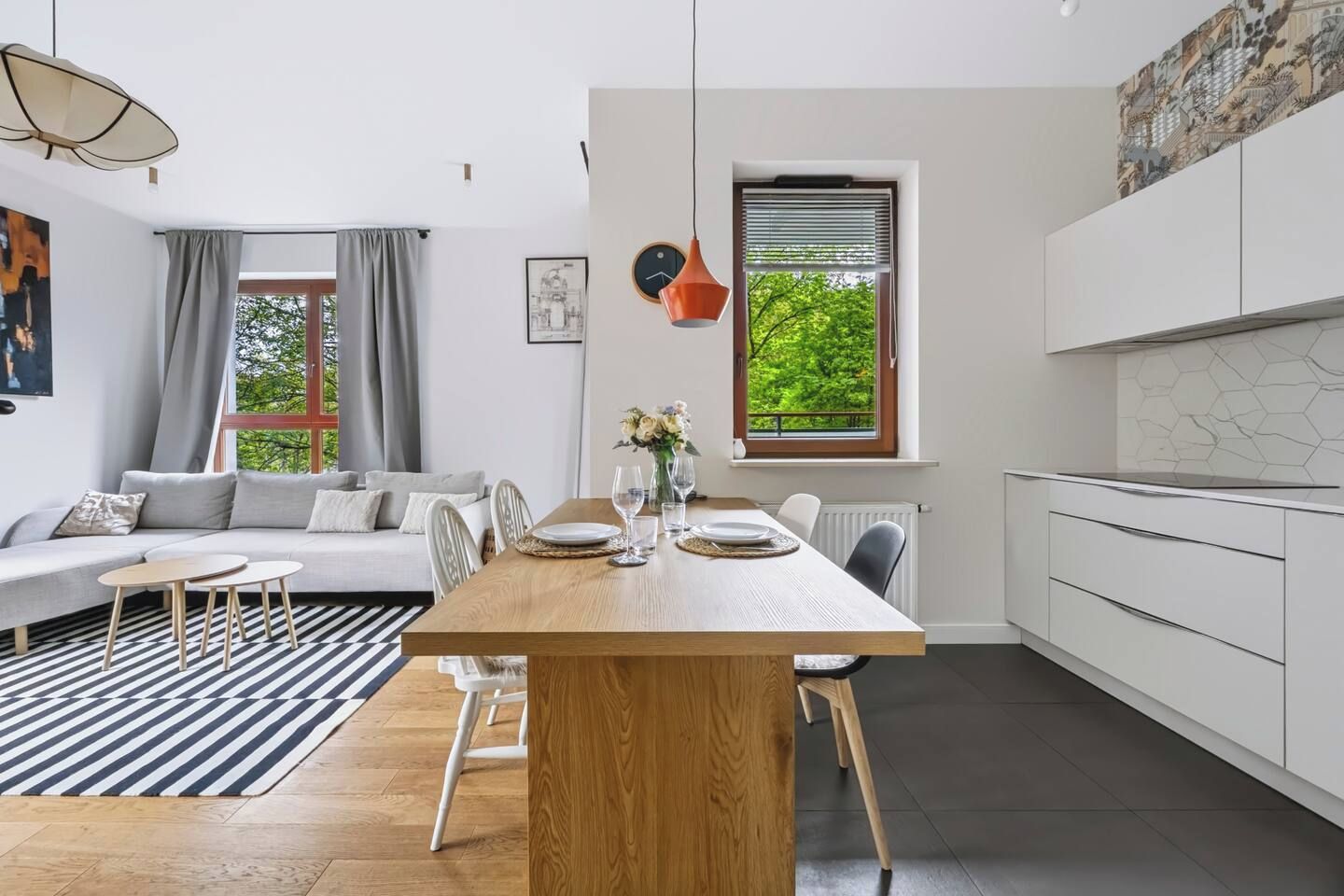 Ett modernt vardagsrum med en soffa, ett matbord och stolar. Det finns ett fönster med persienner, en hängande taklampa och en tavla på väggen.