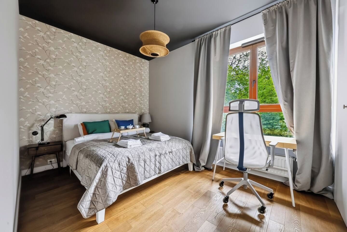 A bedroom with a bed, bedside table, lamp, and a view of greenery through the window.