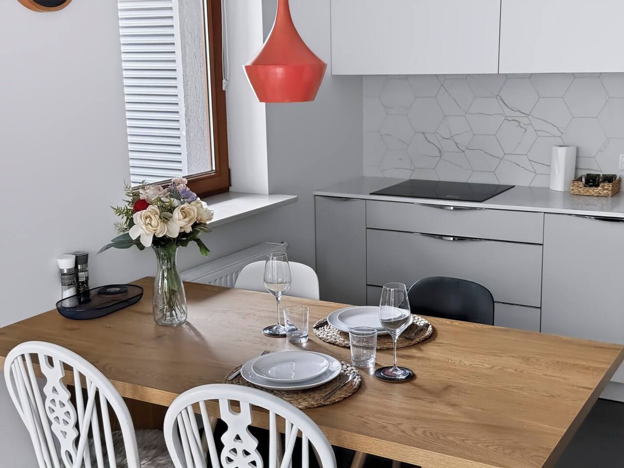 A dining area with a wooden table set for four, white chairs, a vase with flowers, and a modern kitchen in the background.