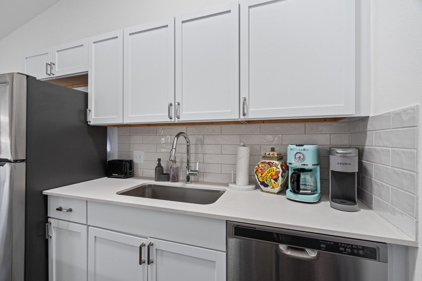 A modern kitchen with white cabinets, a stainless steel refrigerator, a sink, a toaster, a coffee maker, and a dishwasher.