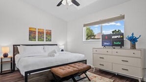 Ein modernes Schlafzimmer mit Bett, Nachttischen, einer Kommode, einem Fernseher, der Streaming-Dienste anzeigt, und einem Fenster mit Blick.