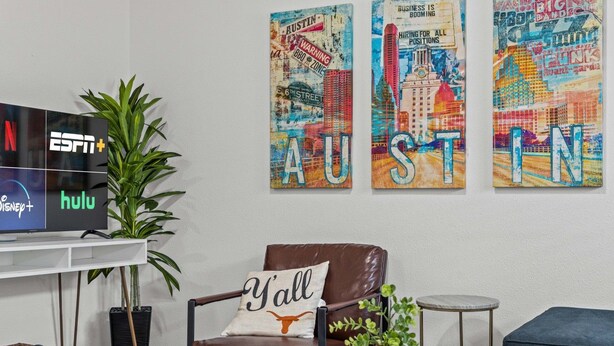 Ein Wohnzimmer mit einem Flachbildfernseher, der die Logos von ESPN+ und Hulu anzeigt, einem braunen Ledersessel mit einem Kissen mit der Aufschrift 'Y'all' und drei farbenfrohen Gemälden mit Austin-Motiven an der Wand.