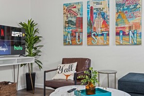 Una sala de estar con un televisor de pantalla plana que muestra los logotipos de ESPN+ y Hulu, un sillón de cuero marrón con un cojín que dice 'Y'all', y tres cuadros coloridos con motivos de Austin en la pared.