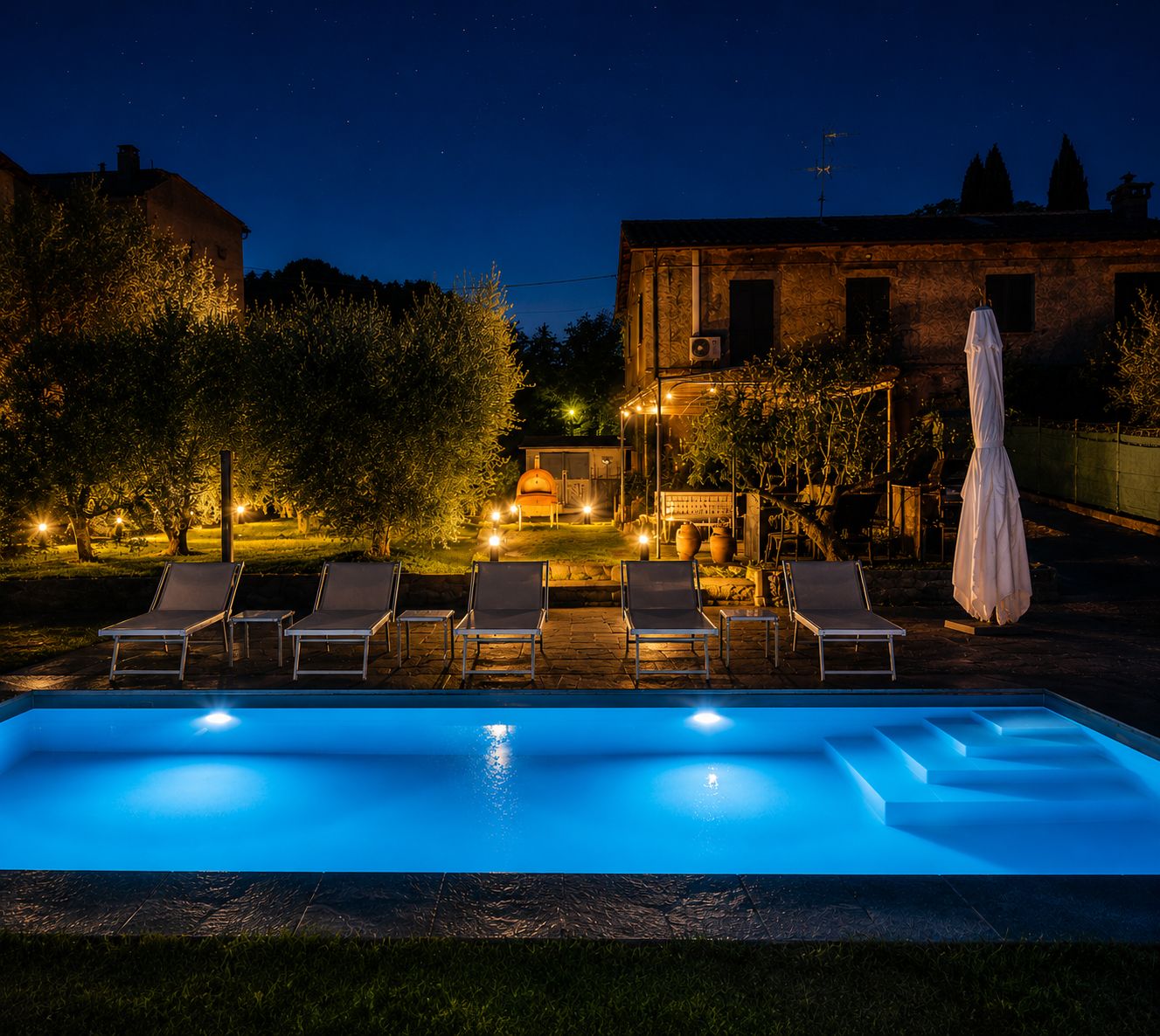 A swimming pool with lounge chairs and a covered umbrella, surrounded by trees and a building in the background.
