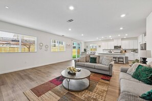 A spacious living room with a grey sofa, a coffee table, and a kitchen in the background. - Sleeps~12, 8-Beds 3-Baths, Backyard, Gameroom, Prime Location (Lincoln)