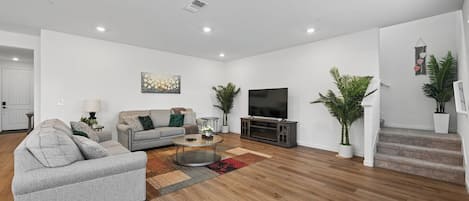 Una sala de estar moderna con un sofá gris, una mesa de centro, un televisor de pantalla plana sobre un mueble de madera y una escalera que conduce a un nivel superior.