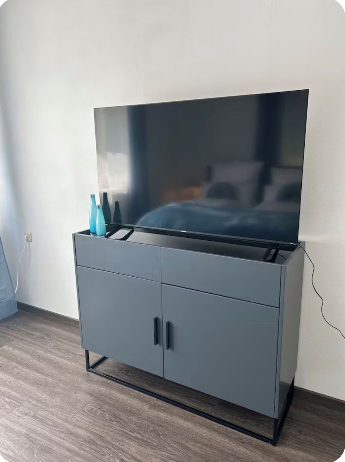 Ein grauer Schrank mit einem Flachbildfernseher darauf, einer Steckdose links und einem Kabel, das mit dem Fernseher verbunden ist.