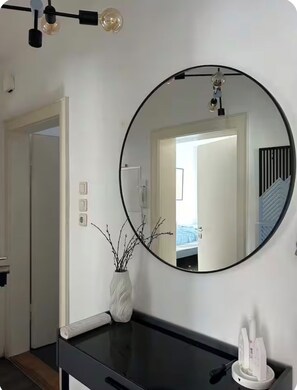 Ein Zimmer mit einem großen runden Spiegel an der Wand, einer weißen Vase mit Zweigen, einer schwarzen Arbeitsplatte und einer Tür, die in einen anderen Raum führt.