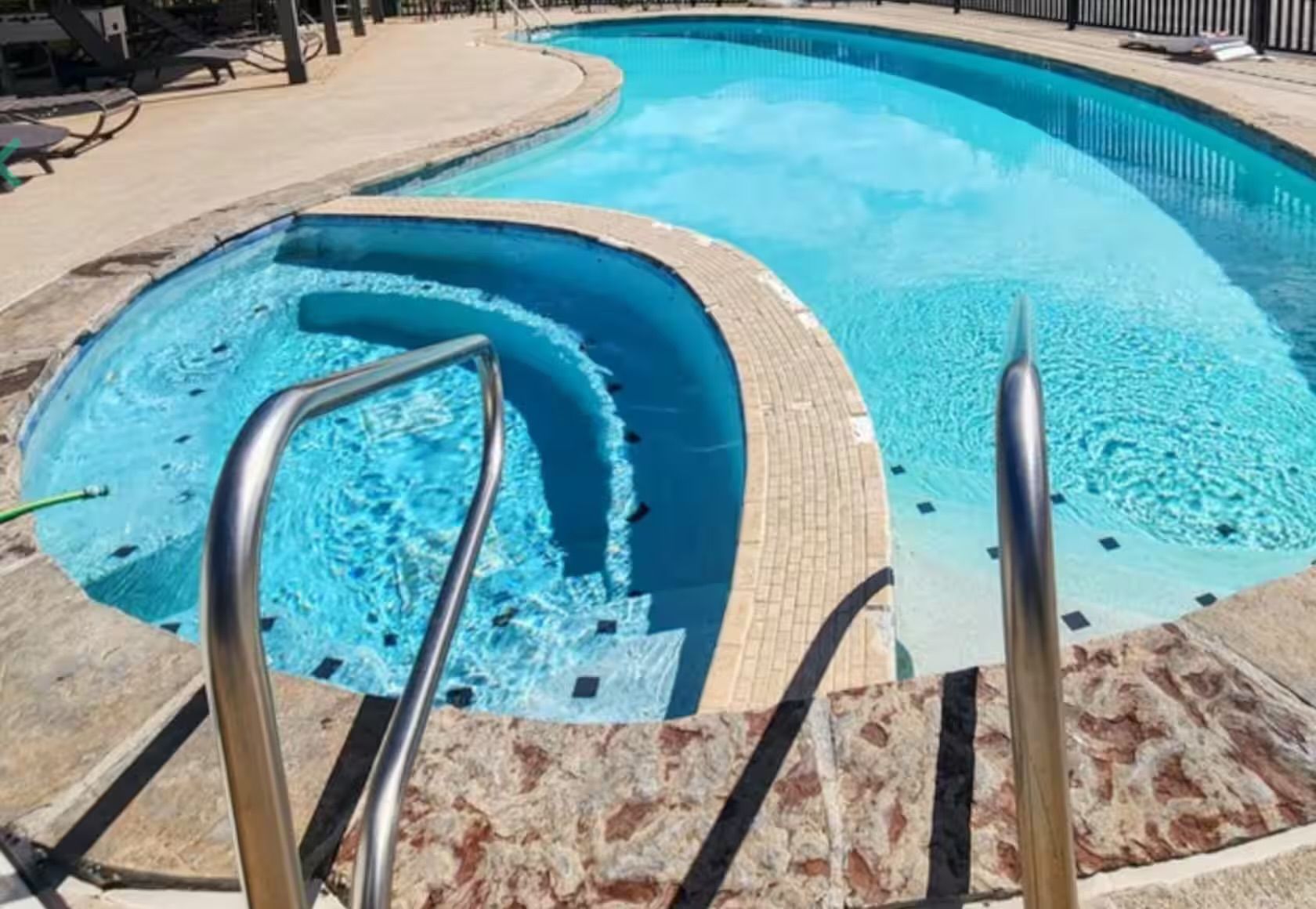 Une piscine au design incurvé, entourée d’une terrasse carrelée et dotée de rampes métalliques.