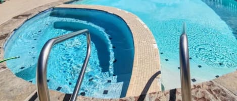 A swimming pool with a curved design, surrounded by a tiled deck and equipped with metal handrails.