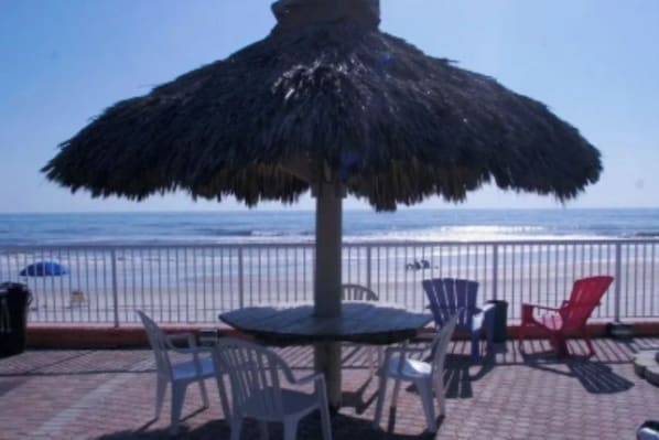 Une terrasse au bord de la plage, agrémentée d’un parasol en chaume, de chaises en plastique blanc et offrant une vue sur l’océan.