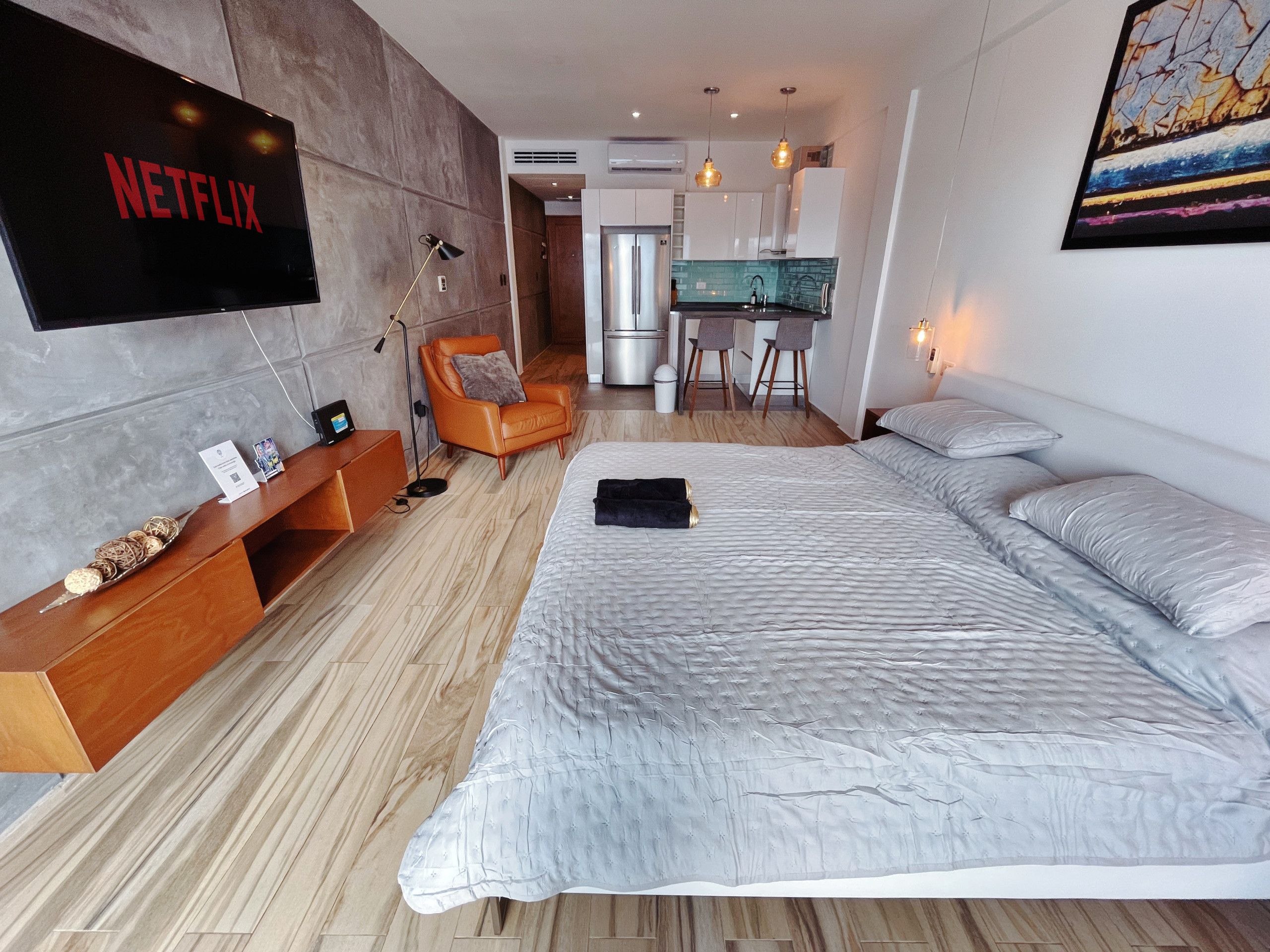 A modern hotel room with a large bed, a desk, a chair, a TV, and a kitchenette.