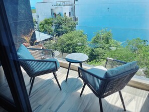 A balcony with two wicker chairs and a small round table, overlooking a cityscape with buildings and trees. - On 5TH AVE! Studio OCEAN VIEW balcony & pool (Playa del Carmen)