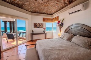 A bedroom with a large bed, a view of the ocean, a balcony with a table and chairs, and a decorative vase.