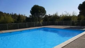 Une piscine rectangulaire dont le bord est recouvert de carreaux, entourée d’une clôture et bordée d’arbres au loin.