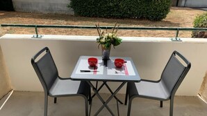 Une terrasse extérieure avec une table et deux chaises, un vase rempli de fleurs et une rambarde en arrière-plan.