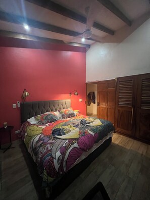 Une chambre à coucher avec une tête de lit capitonnée, une couvre-lit colorée, un ventilateur de plafond et une armoire en bois.