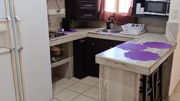 Une cuisine avec des meubles de couleur sombre, un réfrigérateur blanc, un four à micro-ondes et un petit îlot central surmonté de tapis violets.