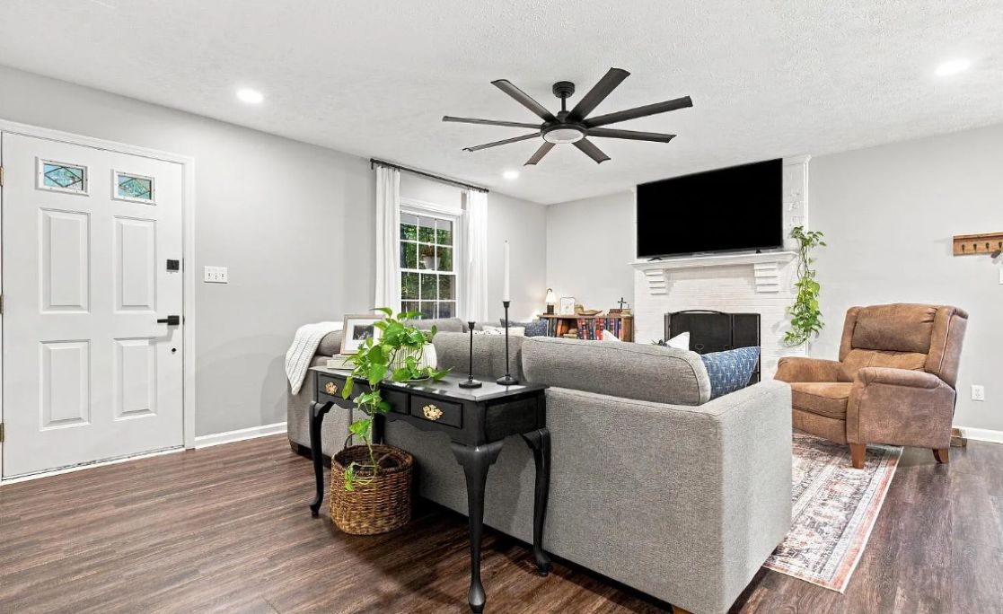 A modern living room with a grey sofa, a brown armchair, a black coffee table, a ceiling fan, and a flat-screen TV mounted on the wall.
