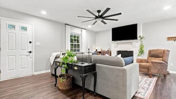 A modern living room with a grey sofa, a brown armchair, a black coffee table, a ceiling fan, and a flat-screen TV mounted on the wall.