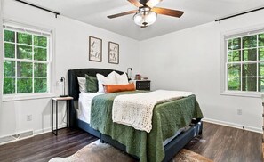 Un dormitorio con cama, mesita de noche, ventilador de techo y dos ventanas con vista a árboles.