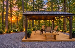 Terraza de madera con pérgola, asientos e iluminación con guirnaldas, en un entorno boscoso.