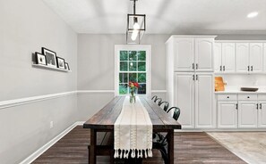 Comedor con mesa de madera, armarios blancos y ventana con vista a follaje.