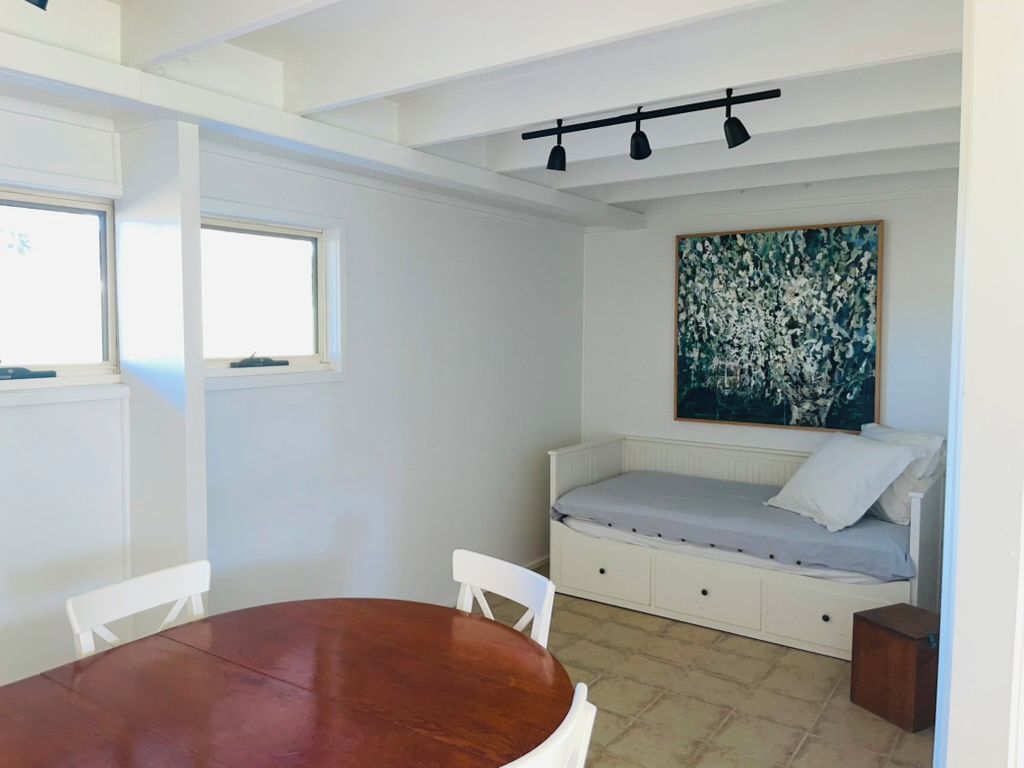 A room with a round wooden table, two white chairs, a white bed with a storage drawer, and a painting on the wall.